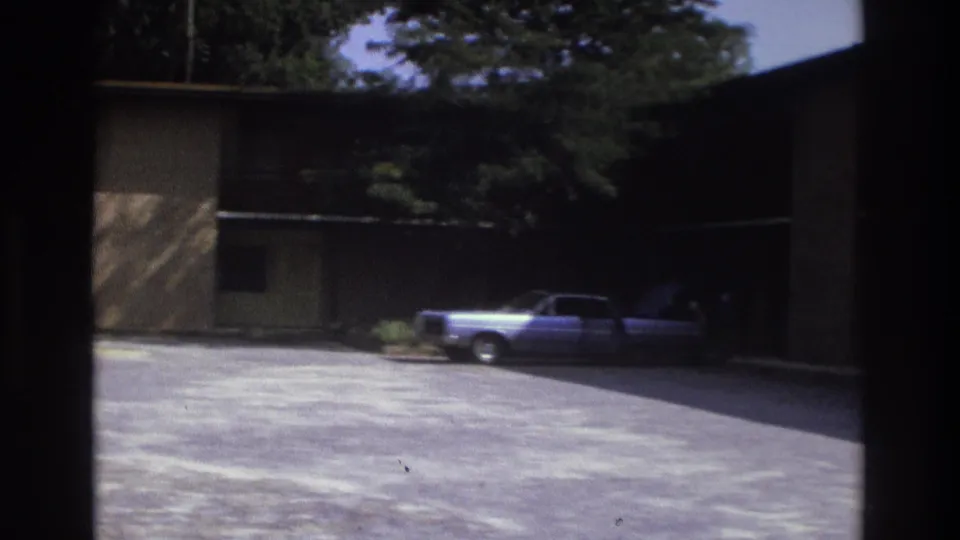 Atlanta 1975 frame of a motel parking lot with a vintage car parked near a shaded two-story building and trees.