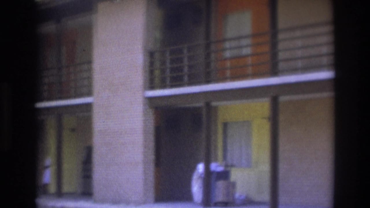 1975 Atlanta home-movie frame showing a two-story brick apartment exterior, balcony rails, walkway posts, and a person by a light-colored car.