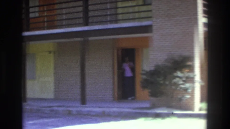 1975 Atlanta frame showing a two-story apartment exterior and a woman in a purple top standing in a ground-floor doorway.