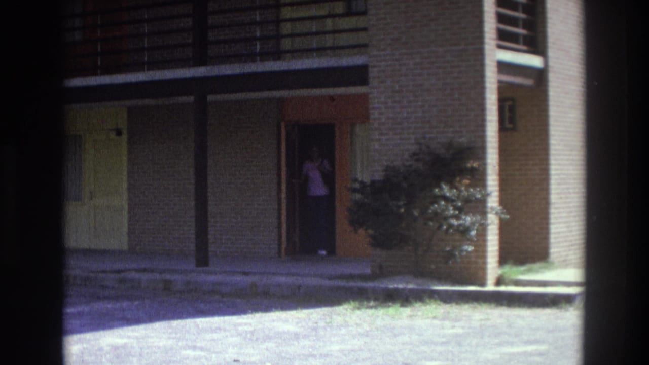 1975 Atlanta 8mm frame showing a woman in a pink top standing inside a brick apartment doorway under a covered exterior walkway.