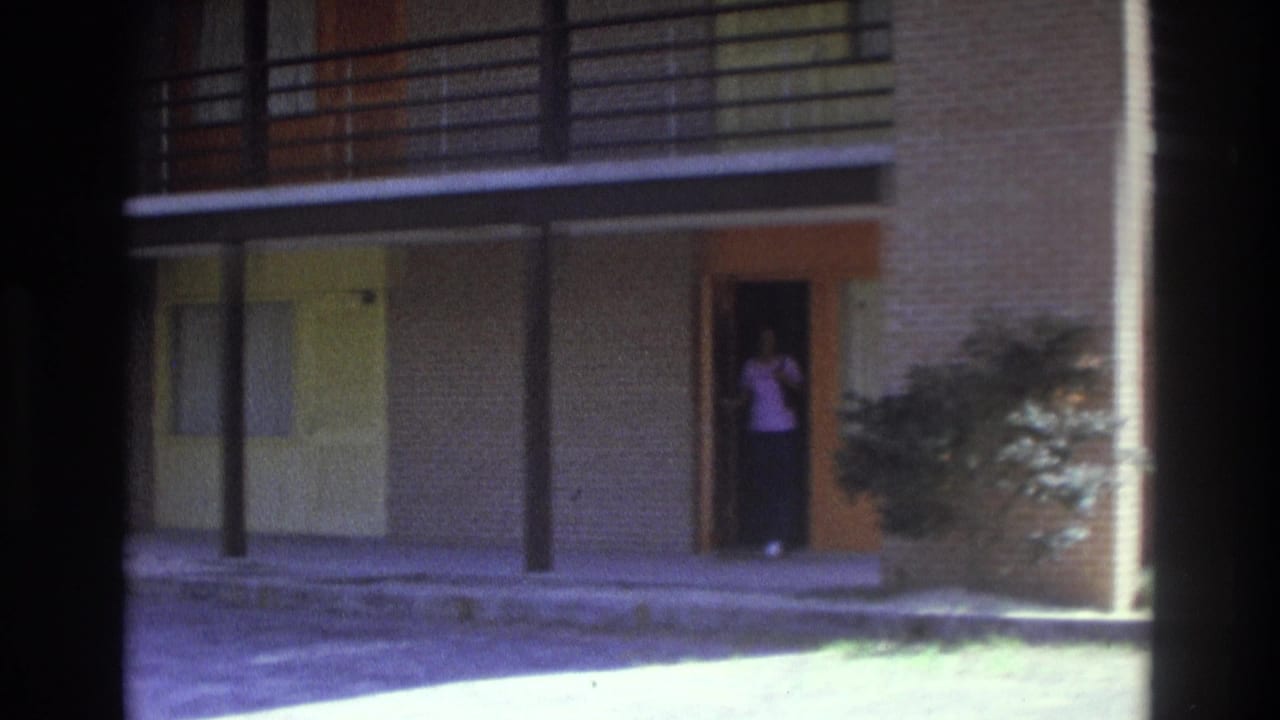 1975 Atlanta home-movie frame with the woman visible at an open orange-trimmed doorway beside shrubbery and covered walkways.