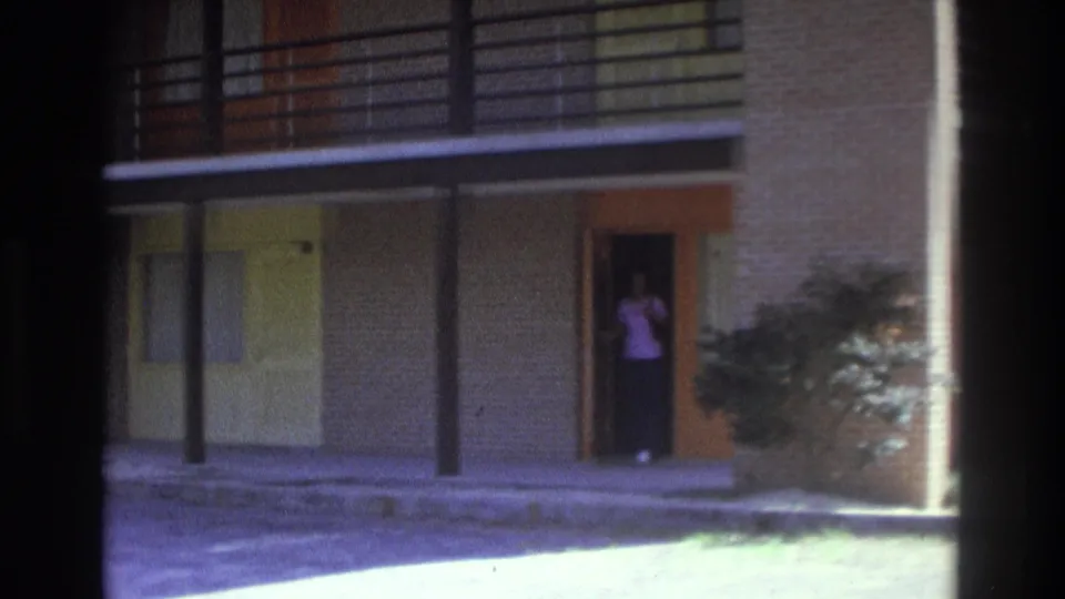 1975 Atlanta home-movie frame with the woman visible at an open orange-trimmed doorway beside shrubbery and covered walkways.