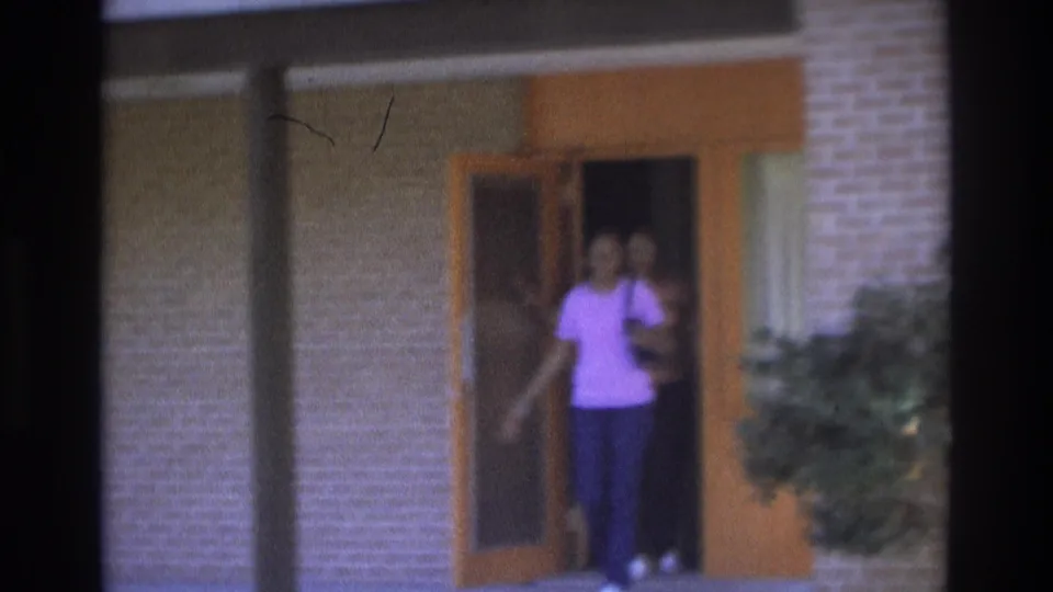 1975 Atlanta frame showing a woman stepping out of a brick apartment doorway, with the open door, walkway posts, and shrub visible.