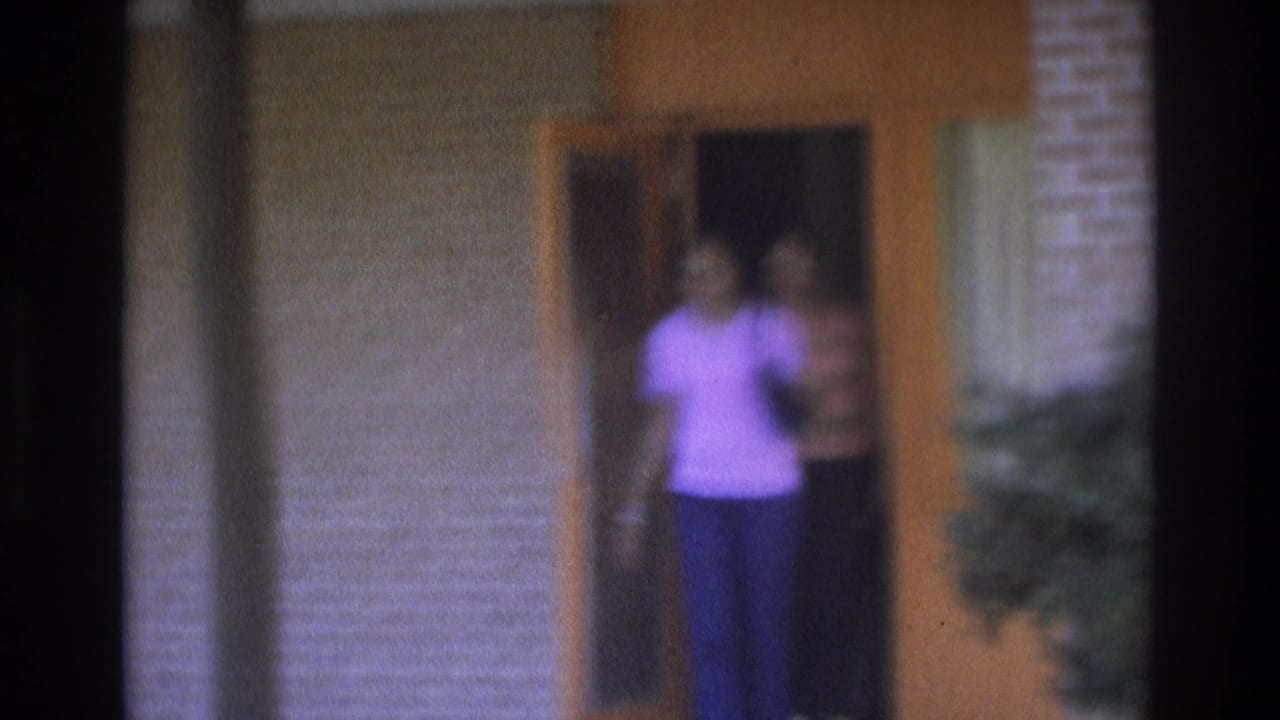 1975 Atlanta home-movie frame showing two women near an orange wooden door on a brick residential exterior.