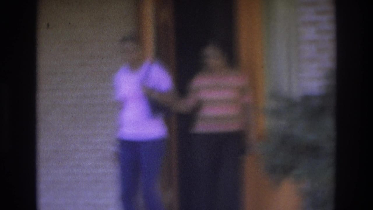 Two women step away from an orange door, one holding an arm out while facing the camera.