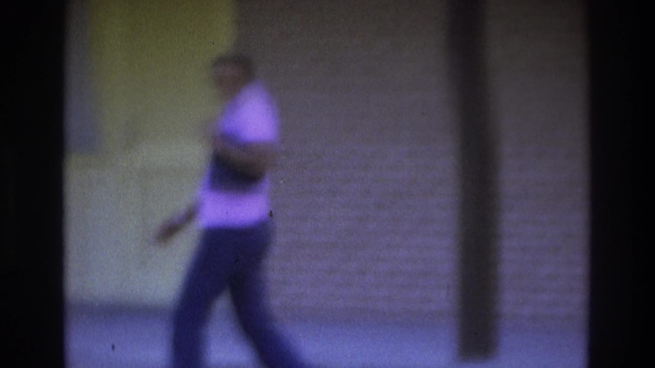 1975 Atlanta 8mm frame showing a woman in a light shirt and dark pants walking along a sidewalk beside yellow and beige walls.