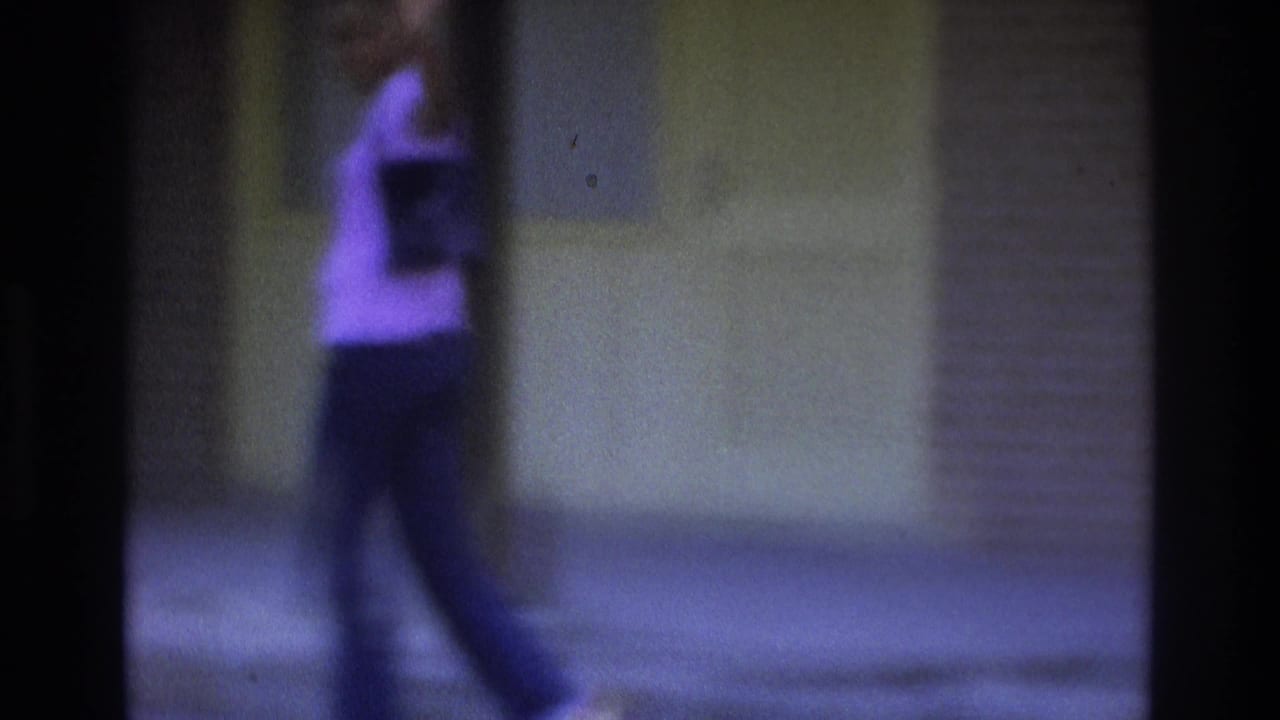 Woman in a light shirt and dark pants walking beside a building wall, partly obscured by a dark post.