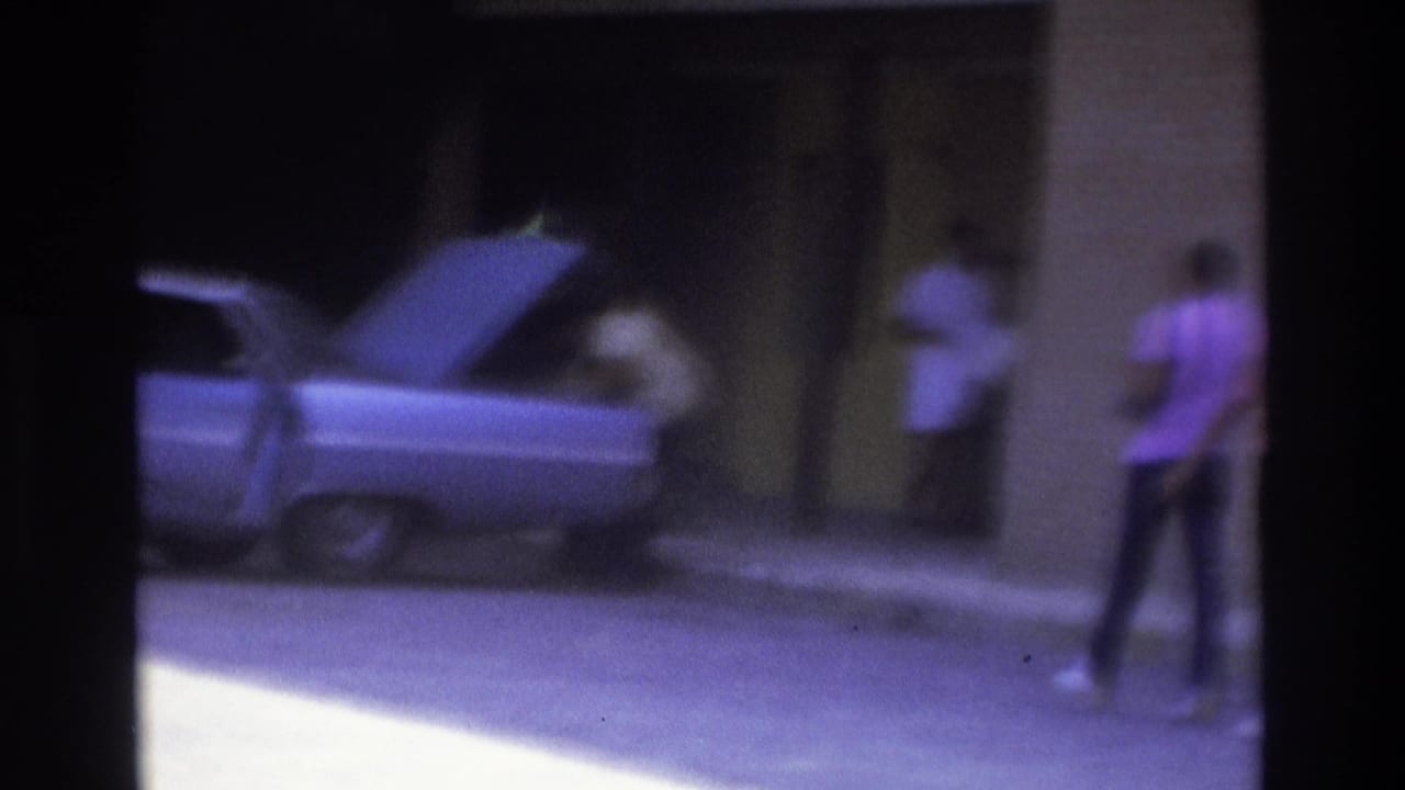 Atlanta 1975 home-movie frame showing a parked car with its trunk open and people near a building entrance.
