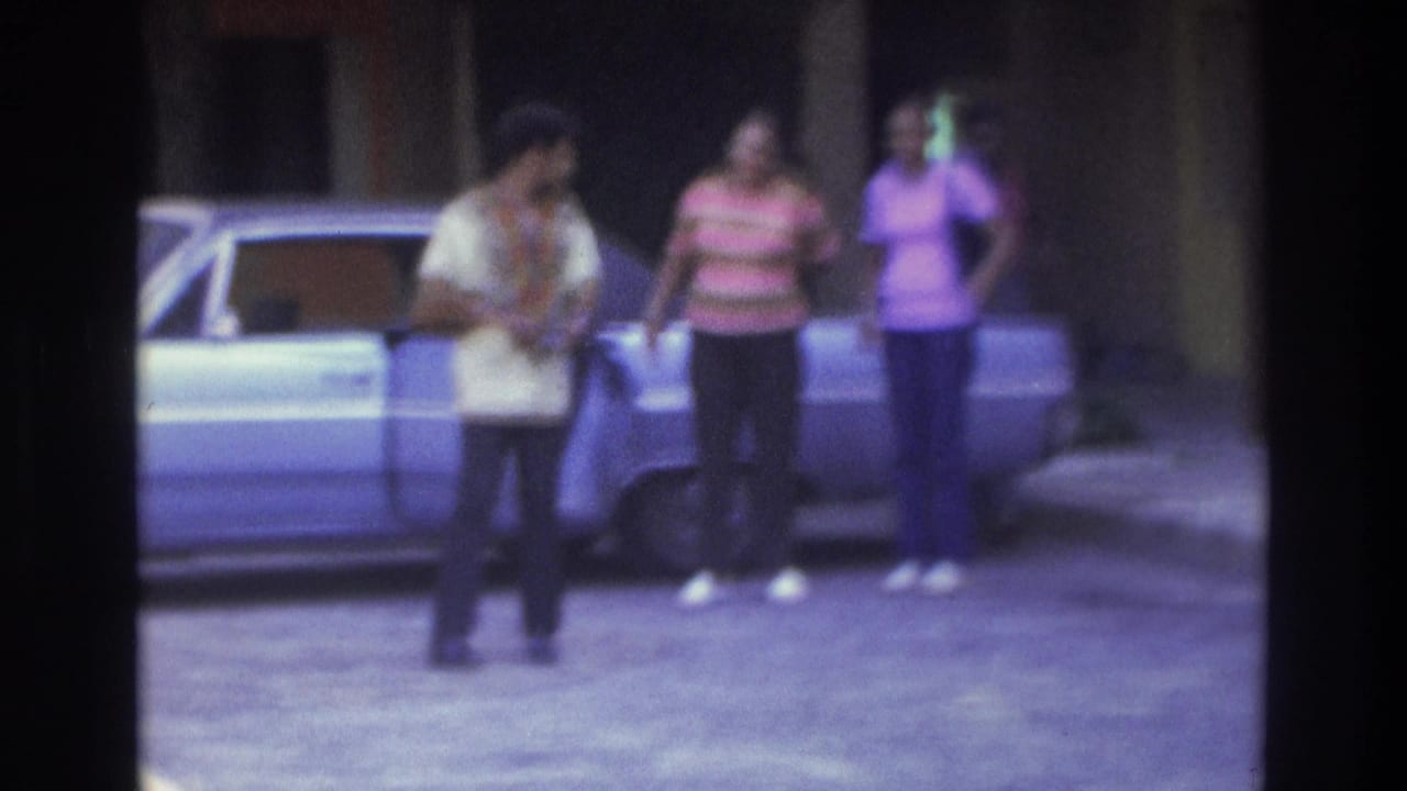 1975 Atlanta home-movie frame showing three adults near a light blue sedan in a sunlit driveway, with soft focus and film color shift.