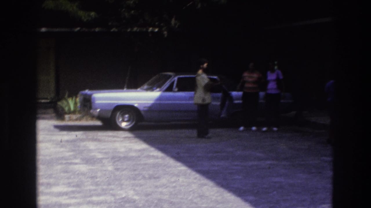 Wide 1975 Atlanta driveway view with several adults near a parked light blue sedan, split between sunlight and deep shade.