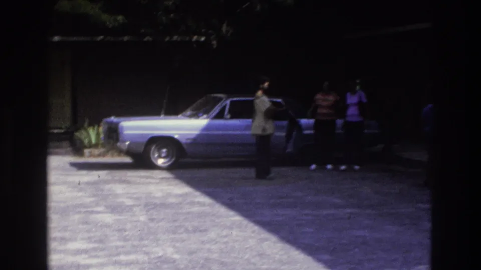 Wide 1975 Atlanta driveway view with several adults near a parked light blue sedan, split between sunlight and deep shade.