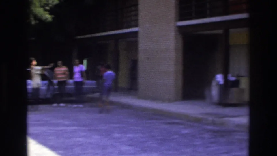 Soft 8mm frame of adults by a light blue sedan outside a residence in Atlanta, with one figure moving across the driveway.