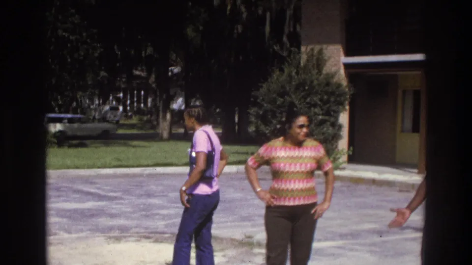 1975 Atlanta home-movie frame showing two adults in casual clothing standing in a bright driveway near a house, lawn, and trees.