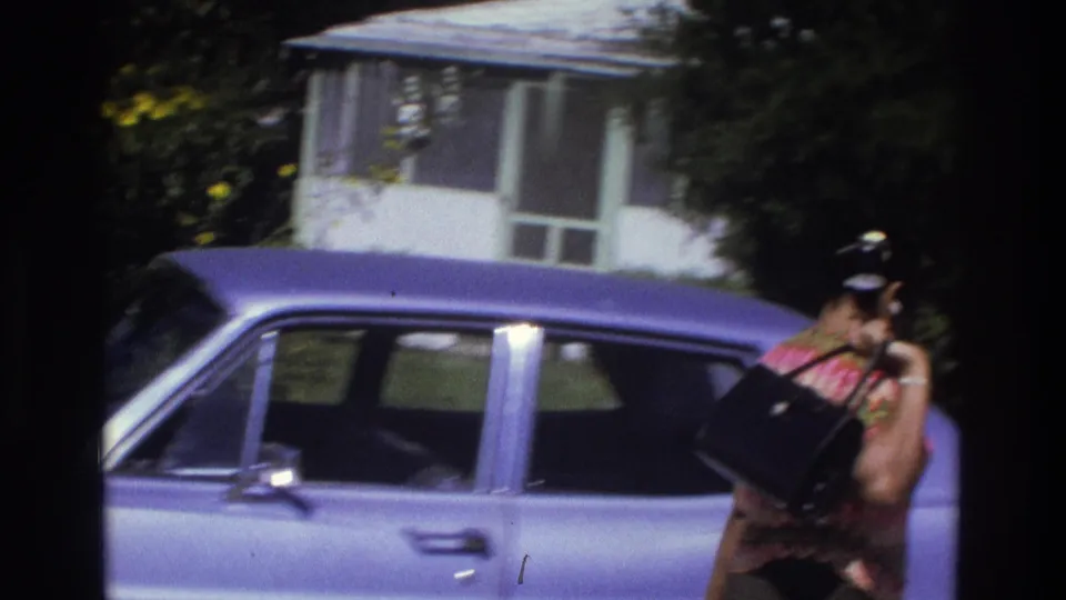 1975 Atlanta home-movie frame showing a woman beside a purple sedan parked near a small house with a screened porch.