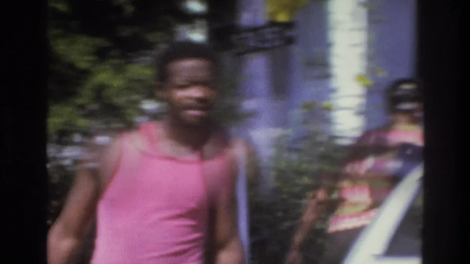 1975 Atlanta frame with a man in a pink tank top near a parked car and another person visible by the house.
