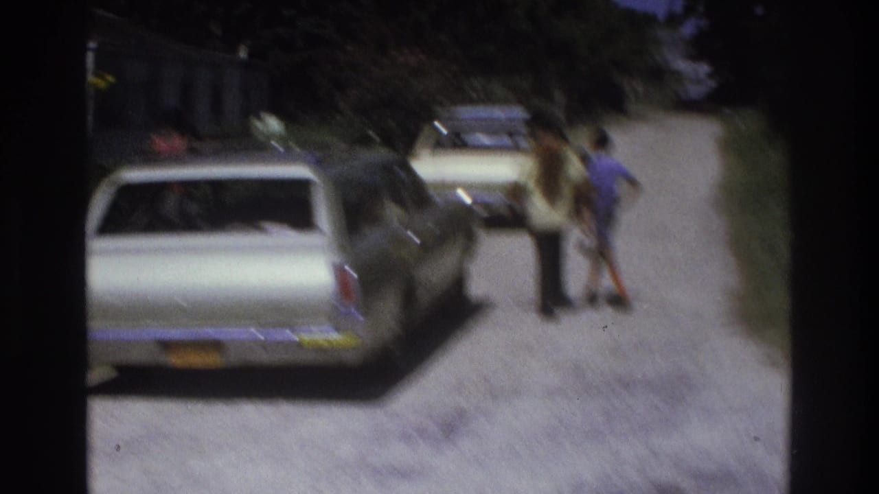 1975 Atlanta home-movie frame showing older cars on a dirt road with two people standing near the parked vehicles.