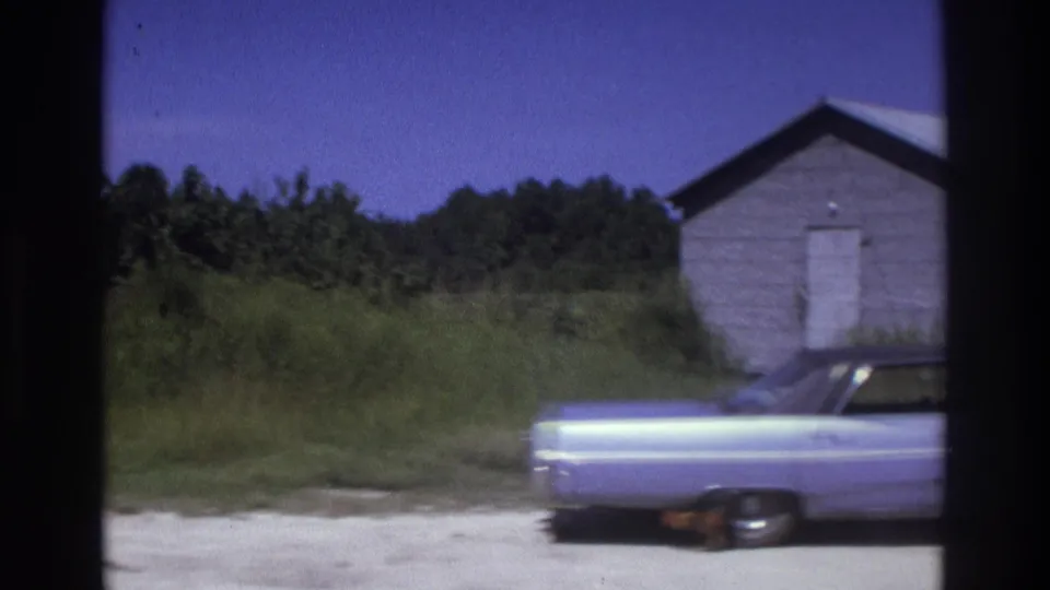 1975 Atlanta-area frame showing a small weathered outbuilding, tall grass, trees, and the rear of a light-colored car.