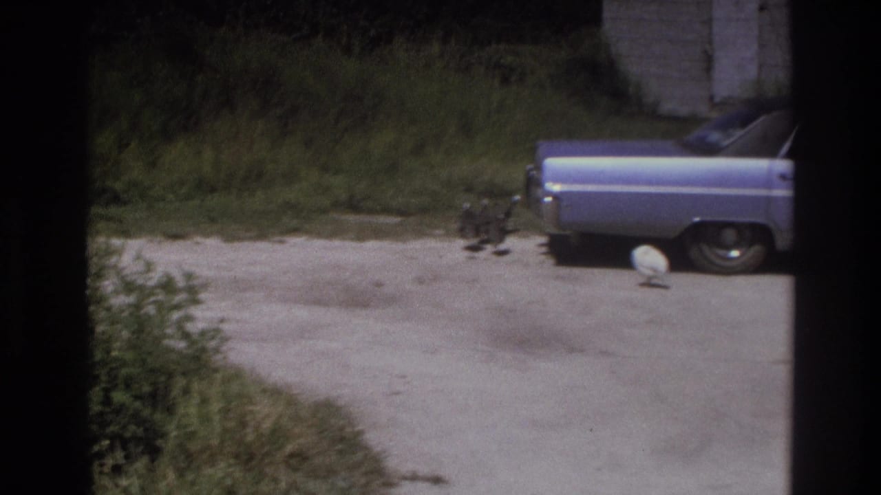 1975 Atlanta-area home-movie frame with a light blue 1970s sedan parked beside a grassy yard and concrete or gravel drive.