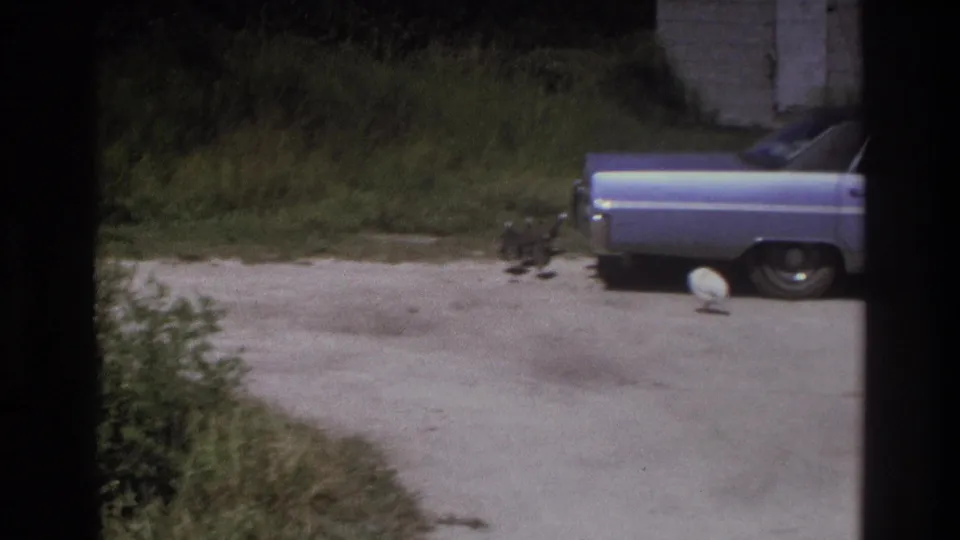 1975 Atlanta-area home-movie frame with a light blue 1970s sedan parked beside a grassy yard and concrete or gravel drive.
