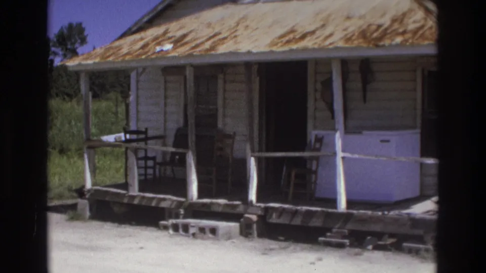 1975 Atlanta-area frame showing a weathered clapboard house porch with wooden chairs, railing, steps, and a rust-stained metal roof.