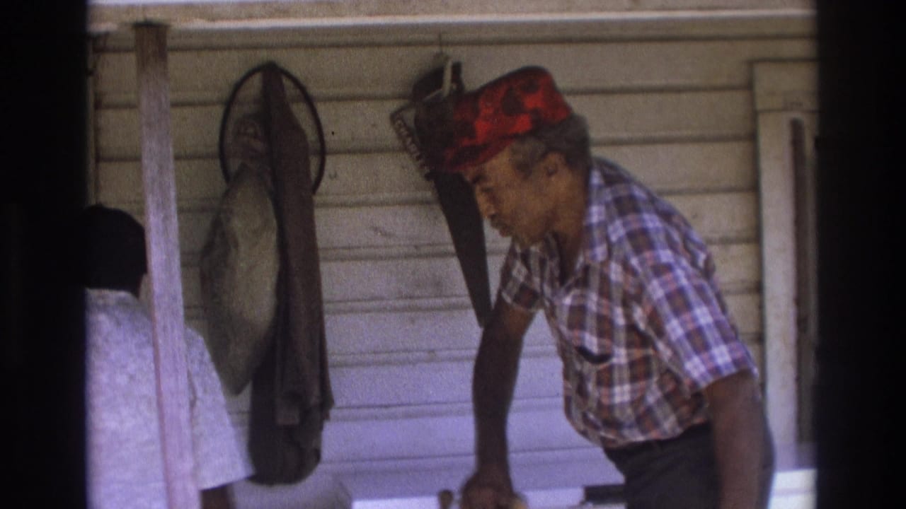 1975 Atlanta home-movie frame of an older Black man on a porch beside a woman, with hanging tools or nets on white siding behind them.