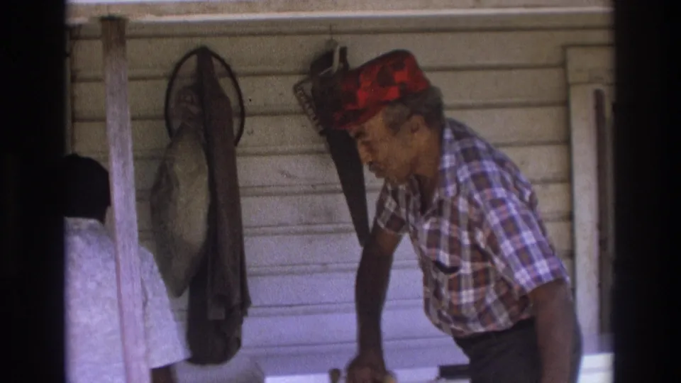 1975 Atlanta home-movie frame of an older Black man on a porch beside a woman, with hanging tools or nets on white siding behind them.