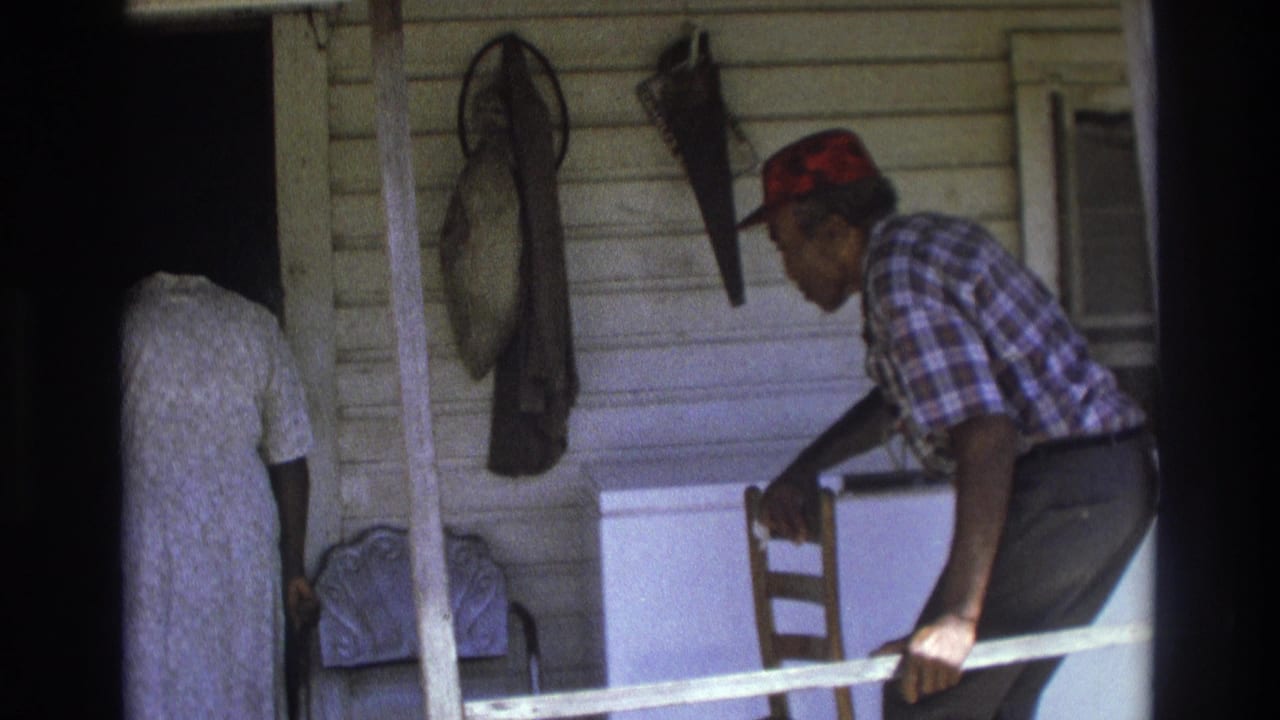 Man in a red cap leans over a porch rail while a woman stands near a doorway.