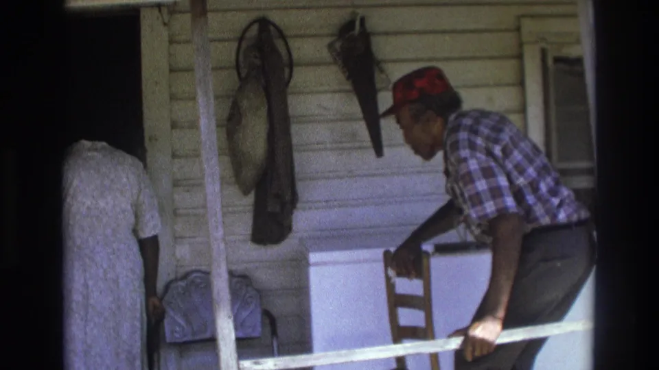 Older Black man in a red cap and plaid shirt leans on a porch rail in 1975 Atlanta, with a woman near the doorway and items on the siding.