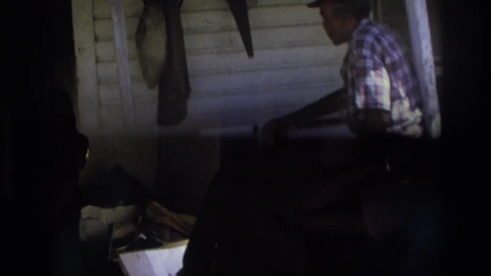 Low-light 8mm frame of the older man on the porch, partly hidden by dark framing, with siding and hanging objects still visible.