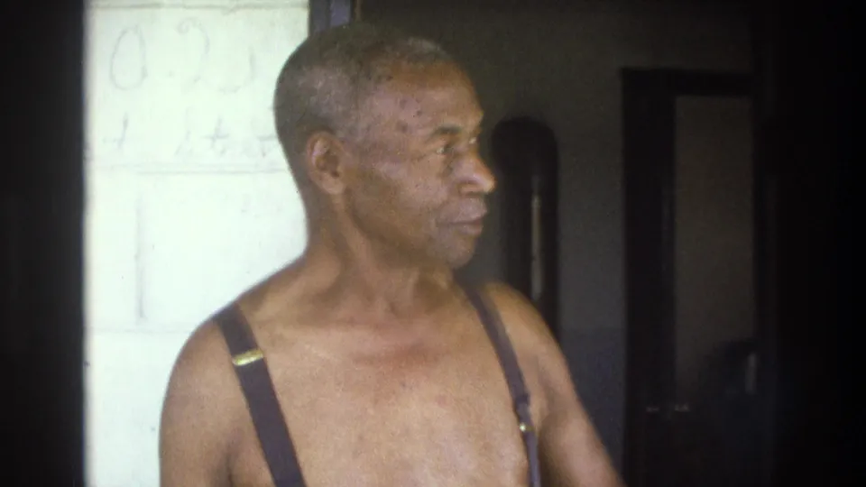 Close interior frame from the 1975 Atlanta home movie showing an older Black man shirtless in suspenders, turned in profile near a pale wall.