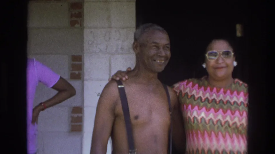 An older Black man in suspenders smiles beside a woman in a colorful patterned top at a doorway in 1975 Atlanta home-movie footage.
