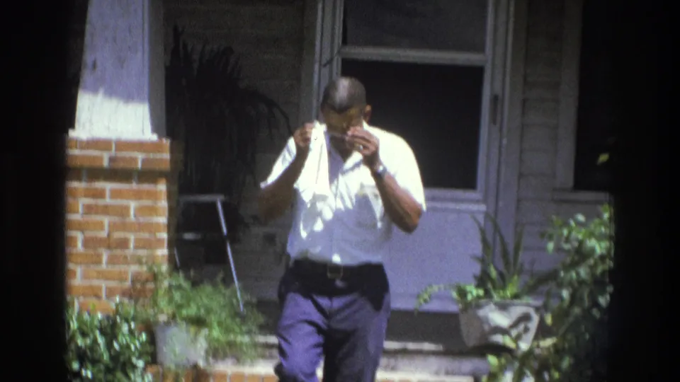 1975 Atlanta home-movie frame showing a man outside a house near brickwork, windows, plants, and a chair, wiping his face.