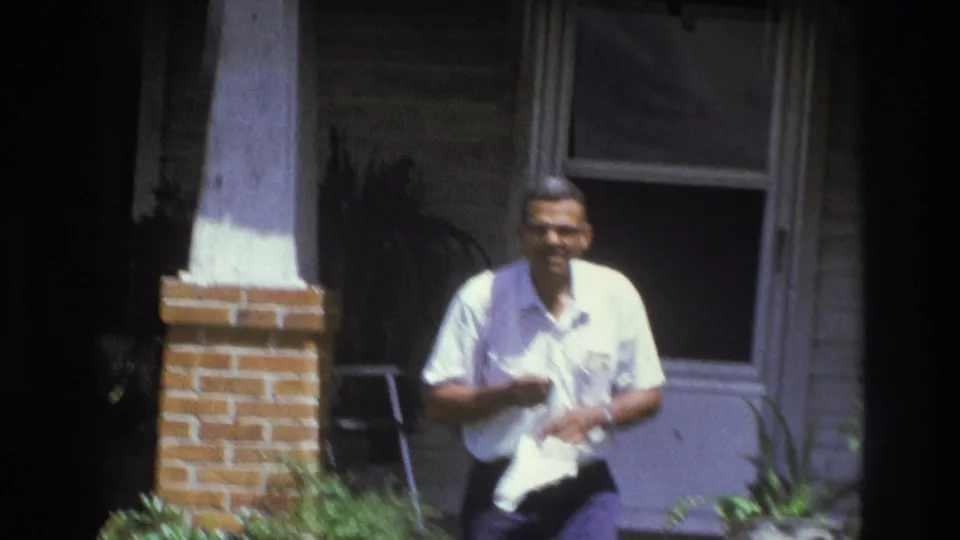 In 1975 Atlanta home-movie footage, an adult man in a white shirt stands on a porch while holding a white towel.