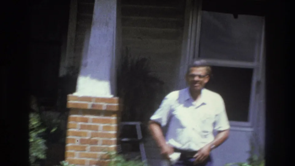 The man smiles toward the camera on a residential porch in Atlanta, with a brick column and window behind him.