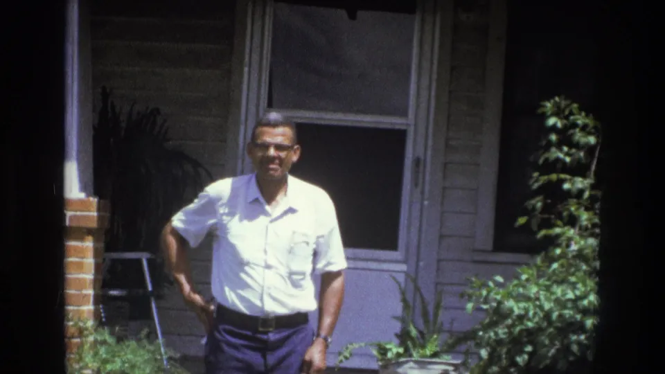 The adult man stands casually on the Atlanta porch, framed by a brick column, window, doorway, and plants.