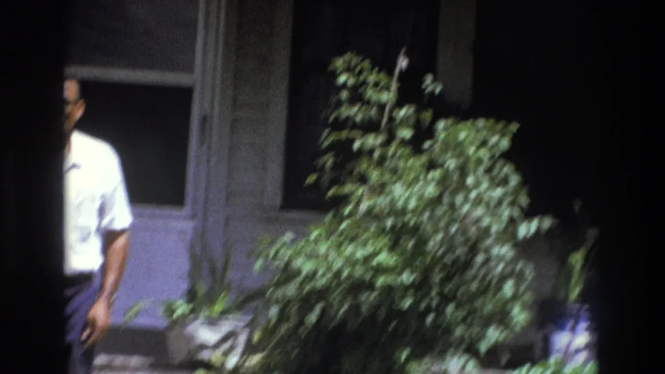 The man is partly hidden by the frame edge and foliage on the residential porch in the 1975 Atlanta home movie.