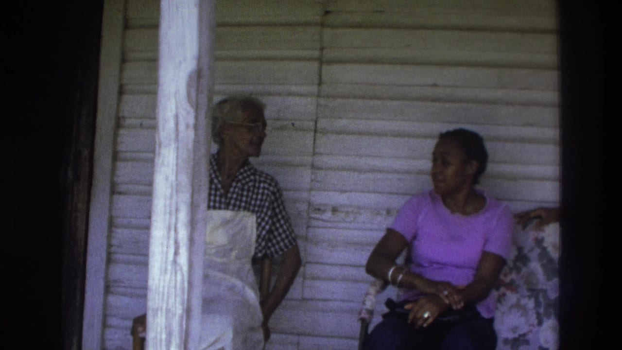 In 1975 Atlanta home-movie footage, two women appear on a porch near white siding, one wearing an apron and one seated in a purple shirt.