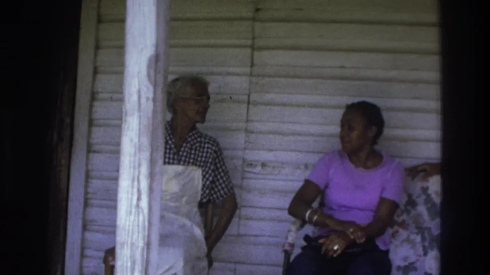 In 1975 Atlanta home-movie footage, two women appear on a porch near white siding, one wearing an apron and one seated in a purple shirt.