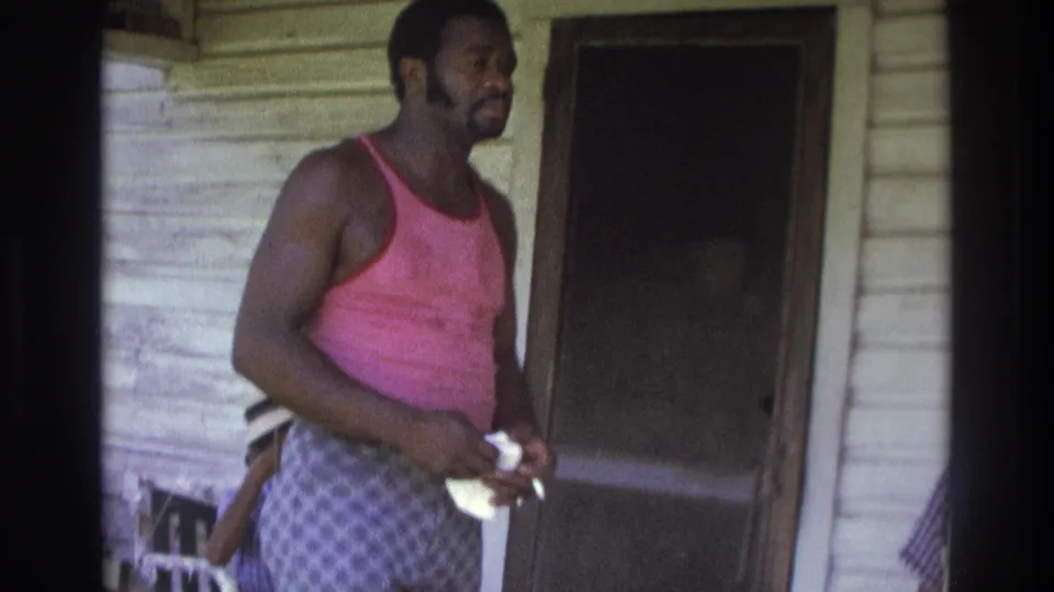 A man in a pink tank top and patterned pants stands on the porch in 1975 Atlanta footage, holding a light-colored object near a screen door.