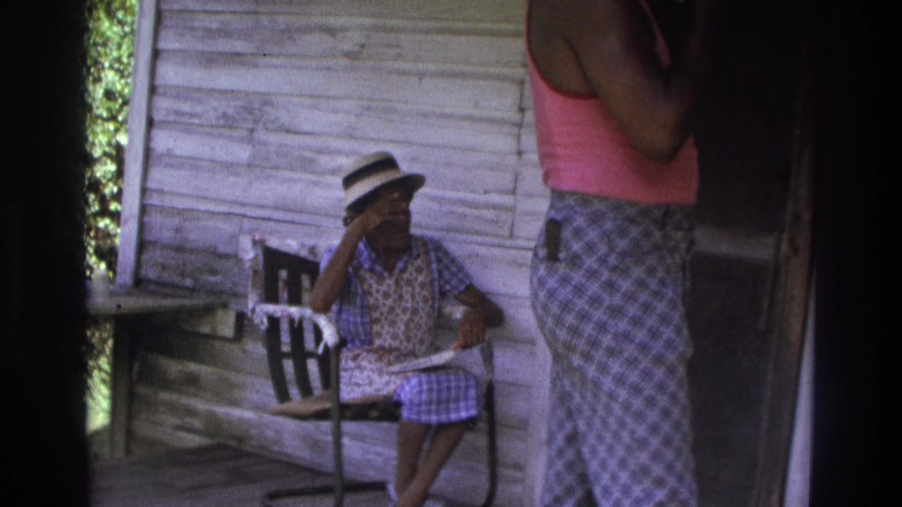 1975 Atlanta home-movie frame showing a woman seated on a porch while another person stands near weathered wood siding and chairs.