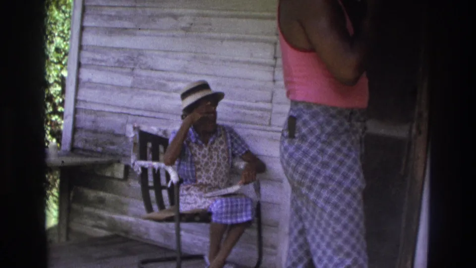 1975 Atlanta home-movie frame showing a woman seated on a porch while another person stands near weathered wood siding and chairs.