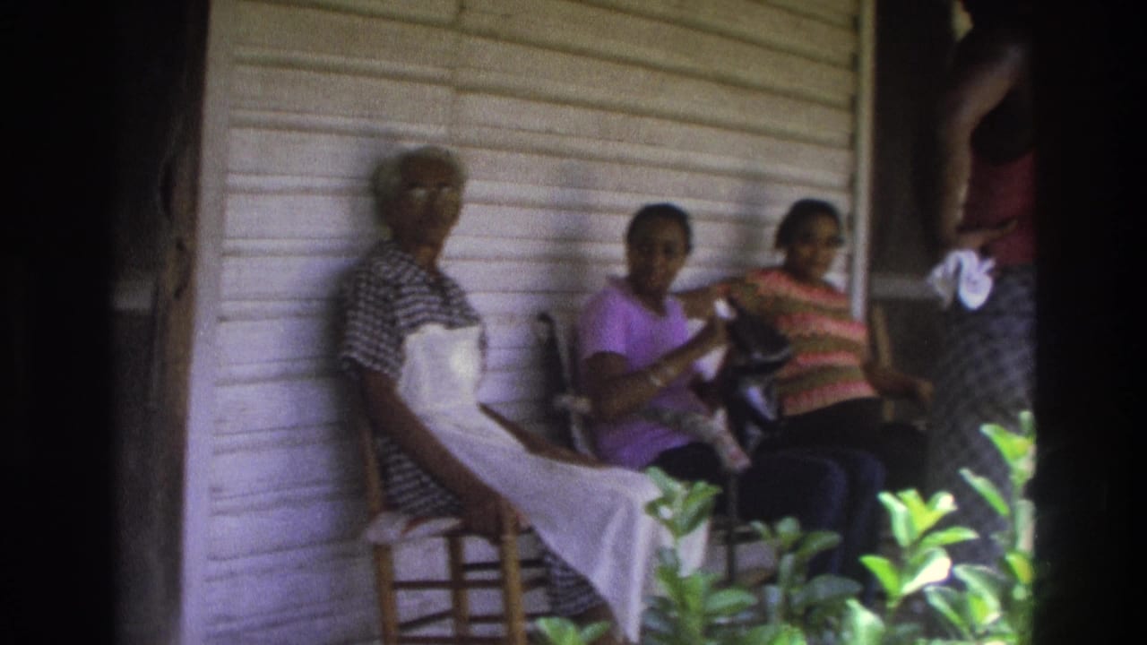 Three women sit on porch chairs beside wood siding while another figure stands partly in frame.