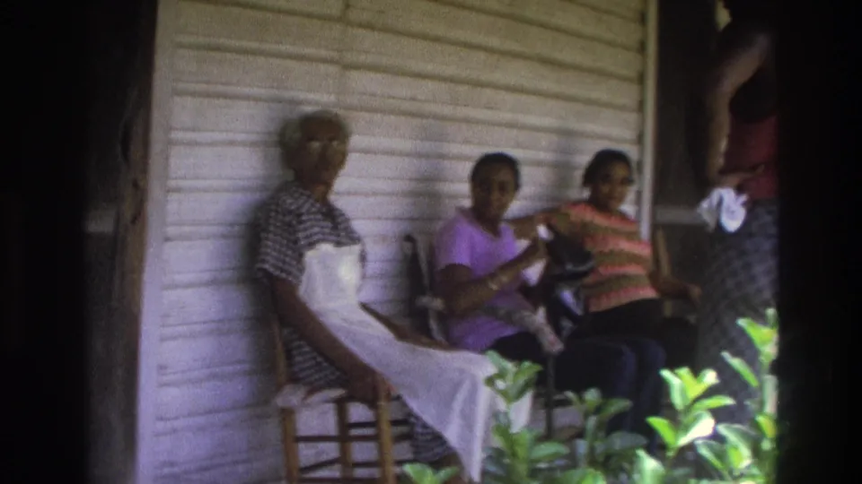 1975 Atlanta porch view with an older woman in an apron seated beside two younger women, with a partial standing figure at the edge.
