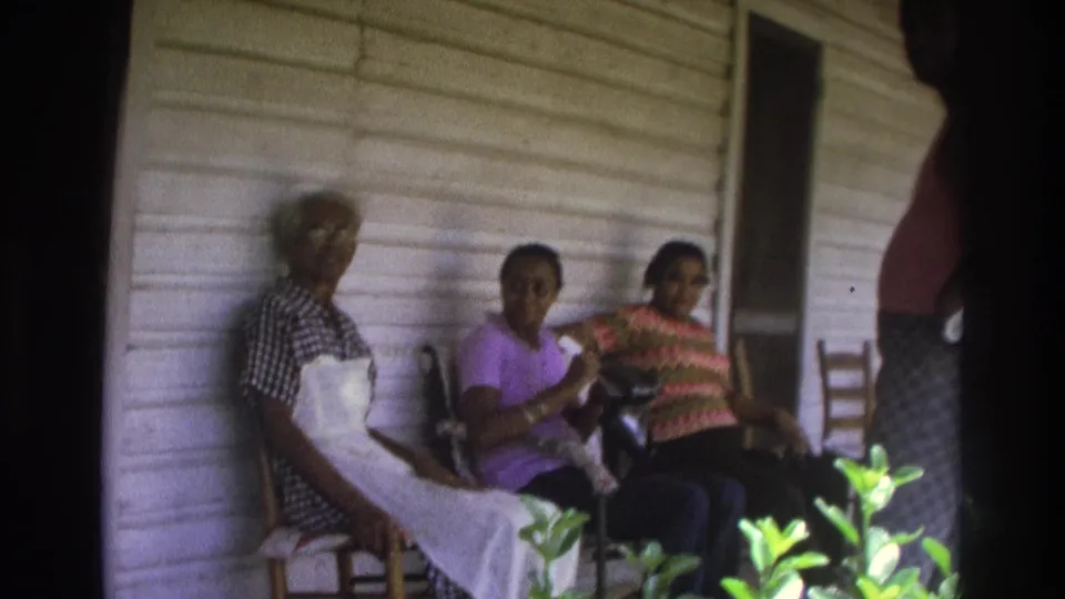 1975 Atlanta home-movie frame of three women seated on a porch beside pale wood siding, with plants and a partial standing figure nearby.