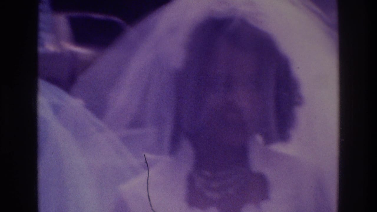 Atlanta 1975 home-movie frame showing a bride in a pale gown near a groom, with guests partially visible and heavy purple film cast.