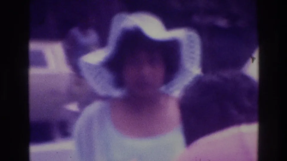 1975 Atlanta wedding frame with a veiled bride among guests, including a wide-brim hat, seen in soft purple-tinted 8mm footage.