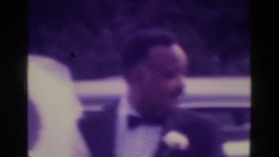 1975 Atlanta home-movie frame showing the groom in a tuxedo with boutonniere beside the bride and surrounding wedding guests.
