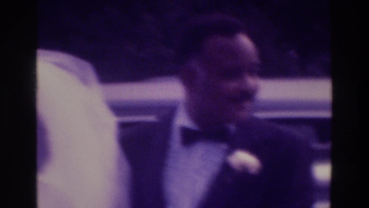 1975 Atlanta home-movie frame showing a groom in a dark tuxedo beside a bride in a white veil near a car.