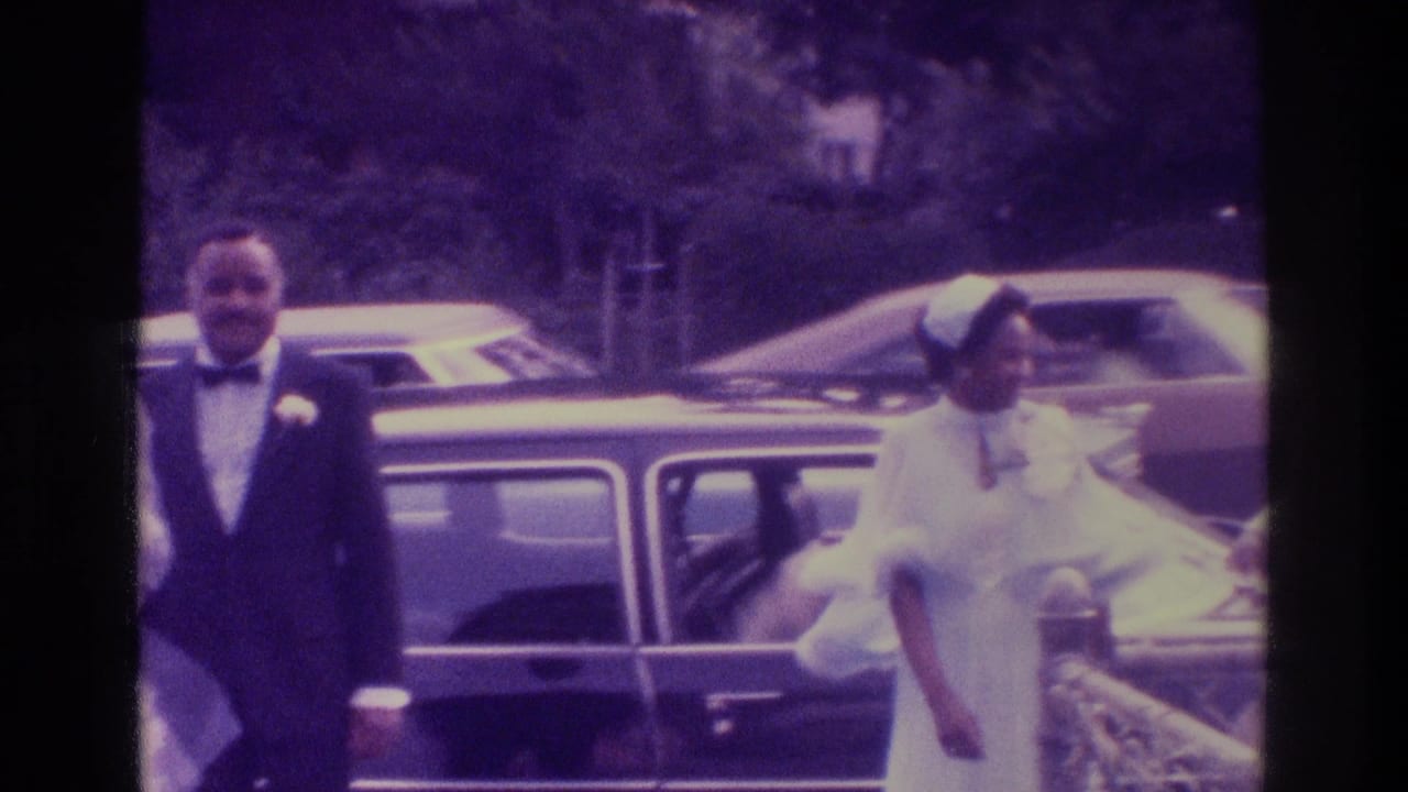 In 1975 Atlanta home-movie footage, a bride in white and groom in a tuxedo stand near a dark classic car outdoors.
