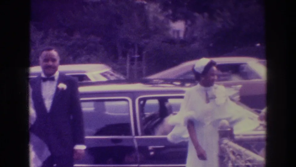 In 1975 Atlanta home-movie footage, a bride in white and groom in a tuxedo stand near a dark classic car outdoors.